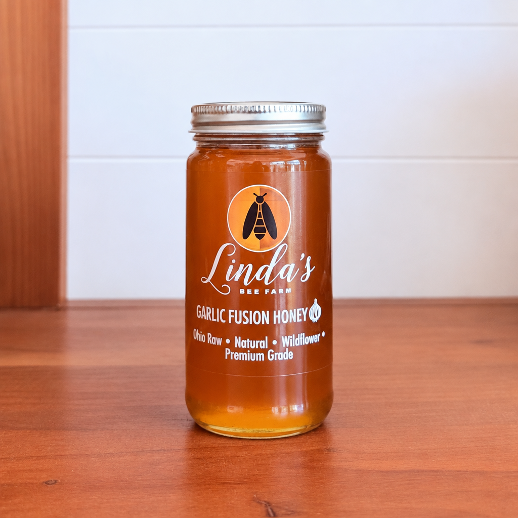A glass jar of Linda's Bee Farm Garlic Fusion Honey sits on a wooden table against a white paneled wall. The jar has a silver metal lid and a white label with a bee logo and product details.